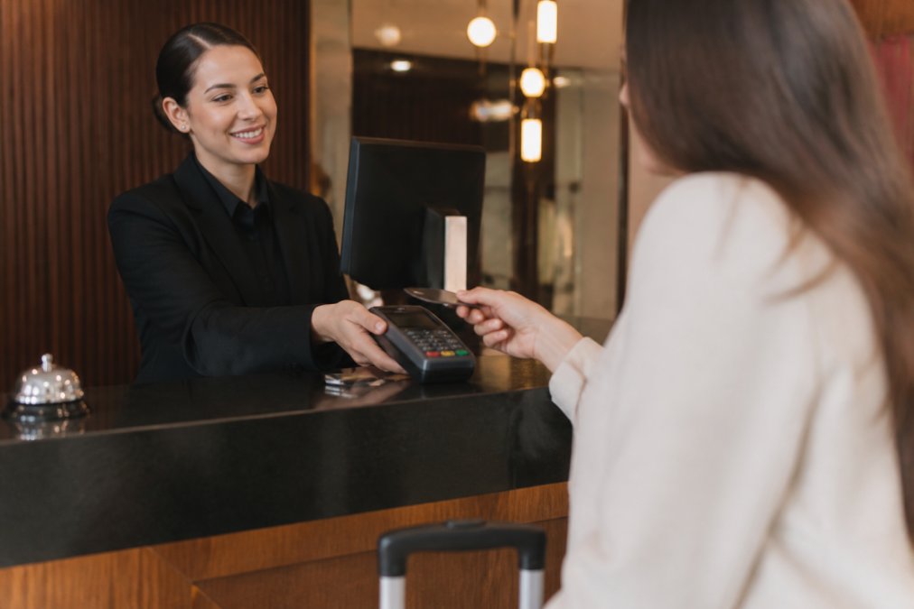 Hotel front desk staff processing a guest payment at a POS terminal, showing the physical interaction with payment processing equipment in a busy hospitality environment