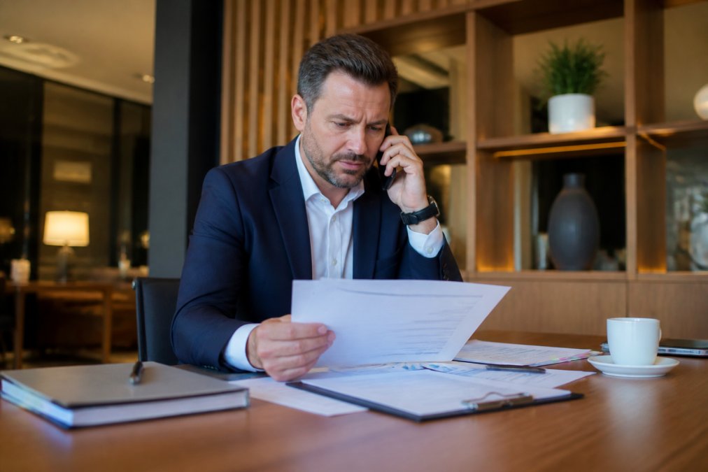 Hotel manager on a phone call in a professional office setting during business hours, appearing focused and serious while handling an urgent situation, natural office lighting, computer and documents visible on desk