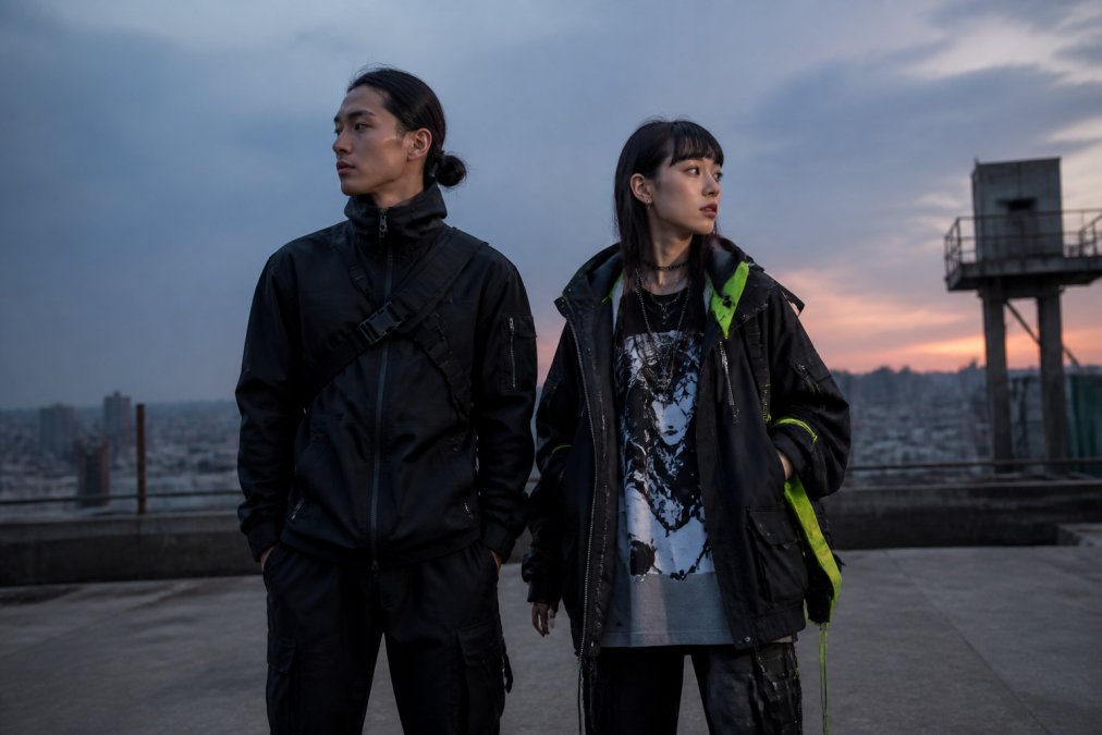 Two people standing side by side in an urban setting at dusk: one in a dark technical jacket with articulated seams, straps, and a tapered silhouette; the other in a layered neon-accented outfit with exposed zippers and a bold graphic tee. Natural street lighting, candid pose.