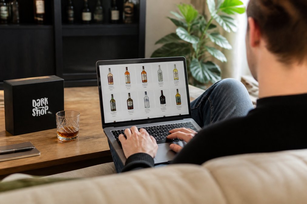 Osoba przeglądająca sklep internetowy na laptopie, różne butelki alkoholu widoczne na ekranie, wygodne otoczenie domowe