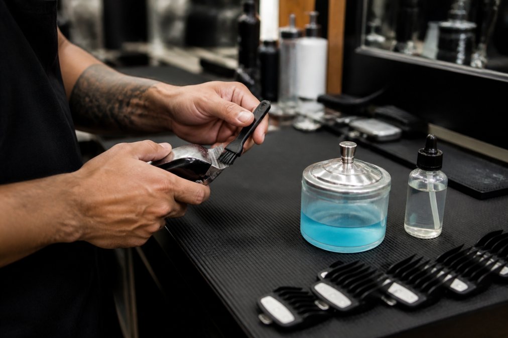 Close-up of a barber's hands using a stiff brush to clean hair buildup from clipper blades at a barber station, with a dip jar and clipper oil visible on the counter