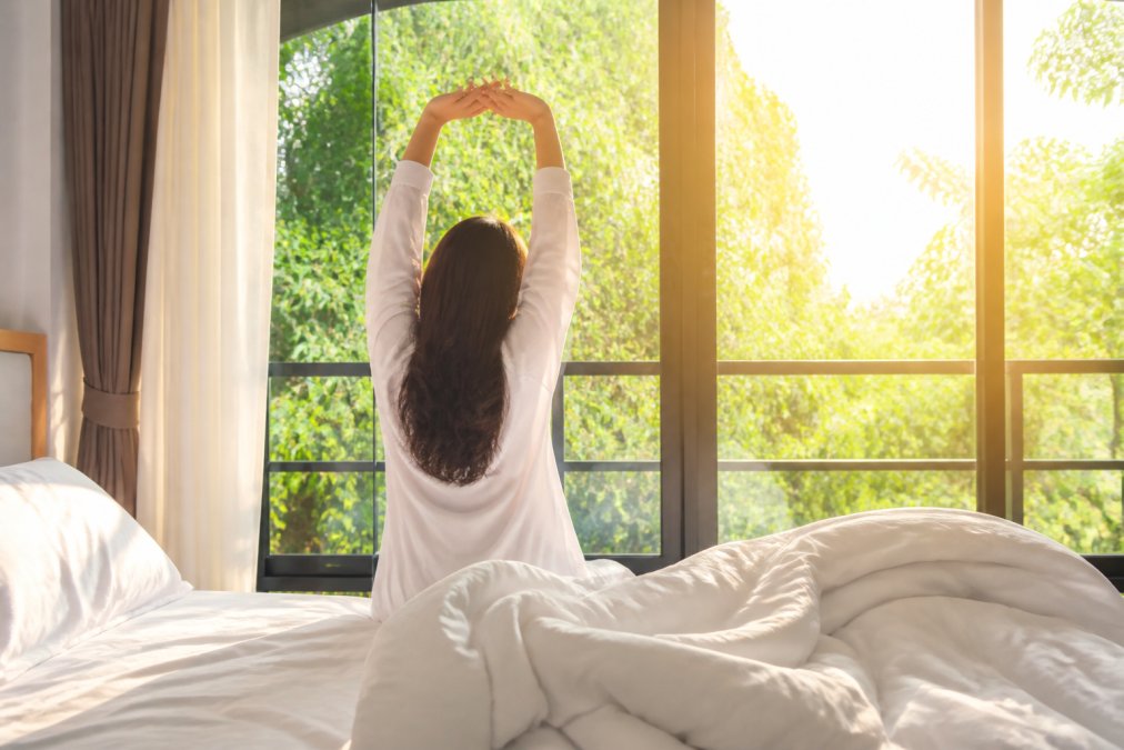 Person waking up in a bright bedroom with natural sunlight streaming through large windows, stretching with arms overhead and a refreshed expression, morning light creating warm tones across the room