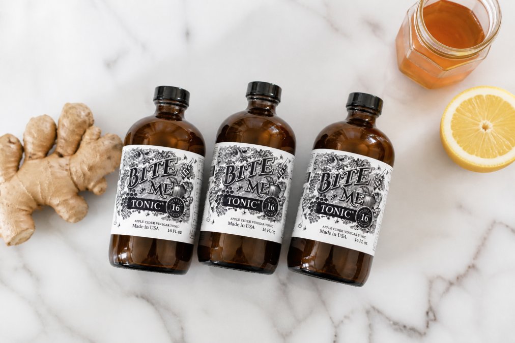 Flat lay of three small glass wellness shot bottles on a white marble surface next to organic ginger root, a small jar of honey, and a halved lemon, with soft natural light