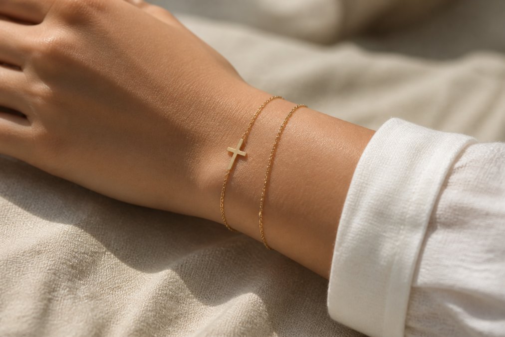 Two stacked dainty gold bracelets with a cross charm resting on a neutral fabric surface in soft light.