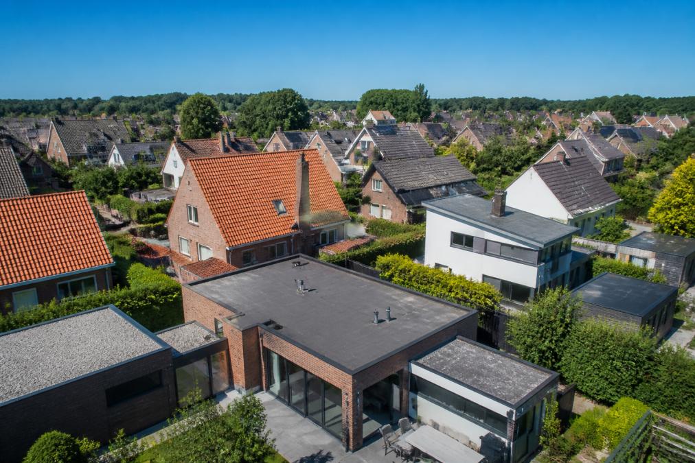 Luchtfoto van residentiële huizen met verschillende dakvormen - zadeldaken met rode dakpannen, moderne platte daken met grijze membranen, en lessenaarsdaken op moderne uitbouwen, tegen een heldere blauwe hemel