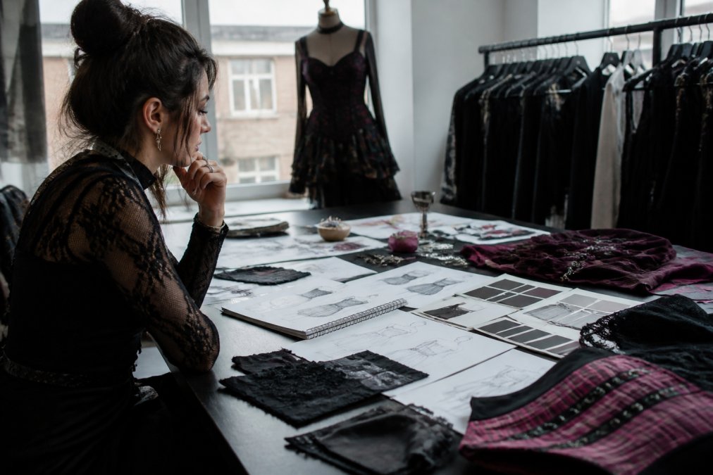 Fashion designer at a modern desk reviewing AI-generated garment concepts displayed on a large monitor, with fabric swatches, physical sketches, and color reference cards spread across the workspace, natural window lighting
