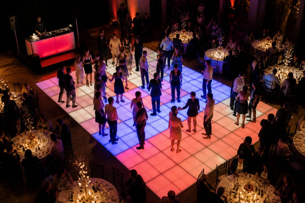 Luchtfoto van een feestzaal met een grote verlichte dansvloer in het midden, omringd door ronde tafels met zittende gasten en een DJ-booth in de hoek, de dansvloer gloeit met kleurrijke LED-verlichting