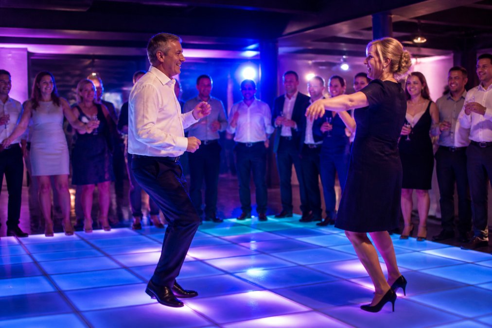 Gastheren dansen op een verlichte dansvloer met dynamische blauwe en paarse verlichting terwijl andere gasten toekijken en genieten op een bedrijfsfeest