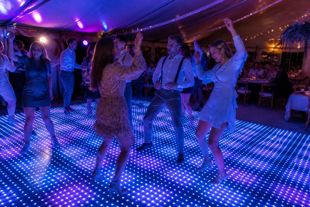 Groep gasten danst energiek op een verlichte LED dansvloer in een feesttent, met kleurrijke blauwe en paarse lichteffecten. Mensen lachen en genieten, met de tent en tafels op de achtergrond