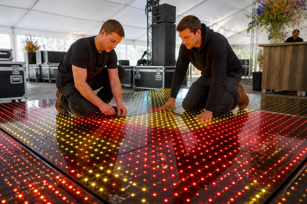 Close-up van een verlichte 100-pixel dansvloer met dynamische rode en gele lichteffecten, waarbij professionele installatietechnici de vloer testen en controleren in een tent
