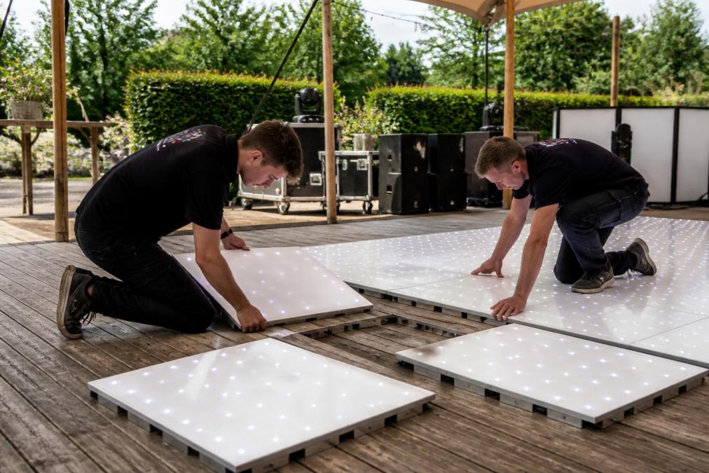 Twee installateurs die samen modulaire dansvloerdelen leggen op een feestlocatie, met zichtbare koppelsystemen en vloerdelen in verschillende stadia van montage, natuurlijke buitenverlichting