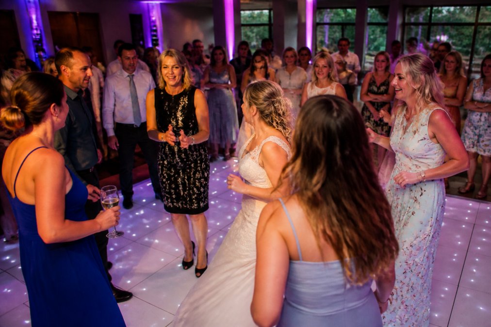 Een groep bruiloftsgasten die dansen op een verlichte dansvloer in een feestzaal, met blije gezichten en energieke poses, terwijl anderen toekijken en genieten van de sfeer