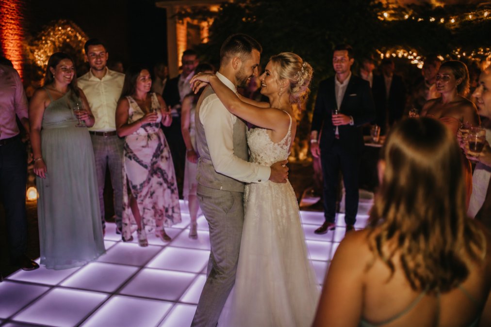 Bruidspaar danst op een verlichte dansvloer met hun gasten, warme sfeerverlichting, lachende gezichten, avondfeest setting