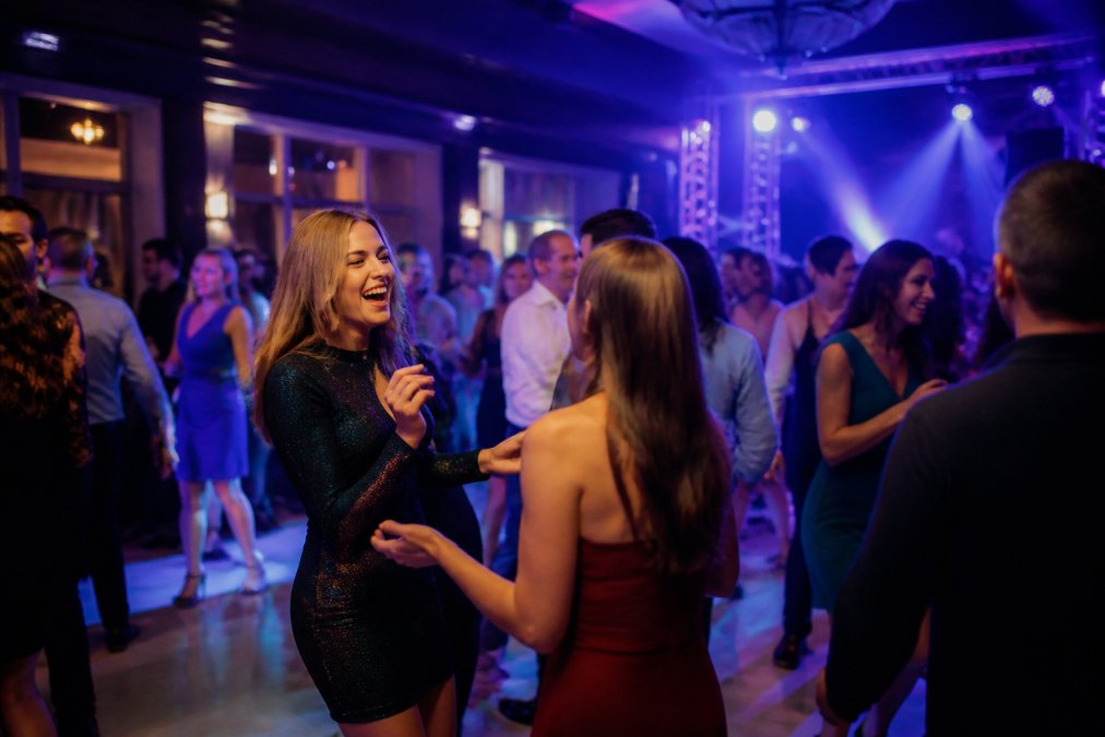 Groep feestgangers van verschillende leeftijden dansend op een verlichte dansvloer met kleurrijke blauwe en paarse verlichting, energiek bewegend, vrolijk gezicht, moderne feestzaal setting