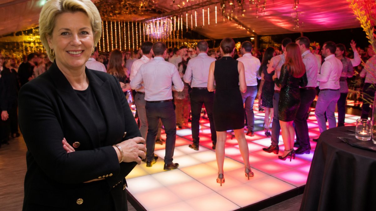 Petra Keizer standing confidently in front of a vibrant illuminated dance floor at an evening event, with guests dancing in the background and warm lighting casting a festive glow across the venue