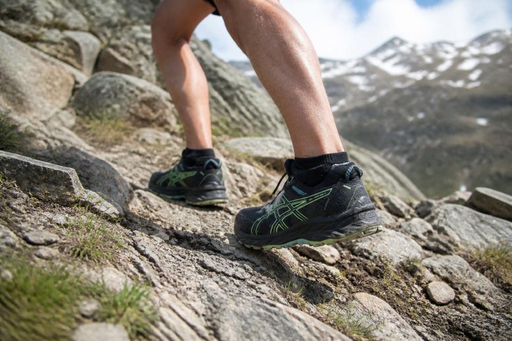 Trail runner in motion on rocky mountain terrain, feet visible navigating sharp rocks and loose gravel, showing athletic stride with moisture-wicking socks in action