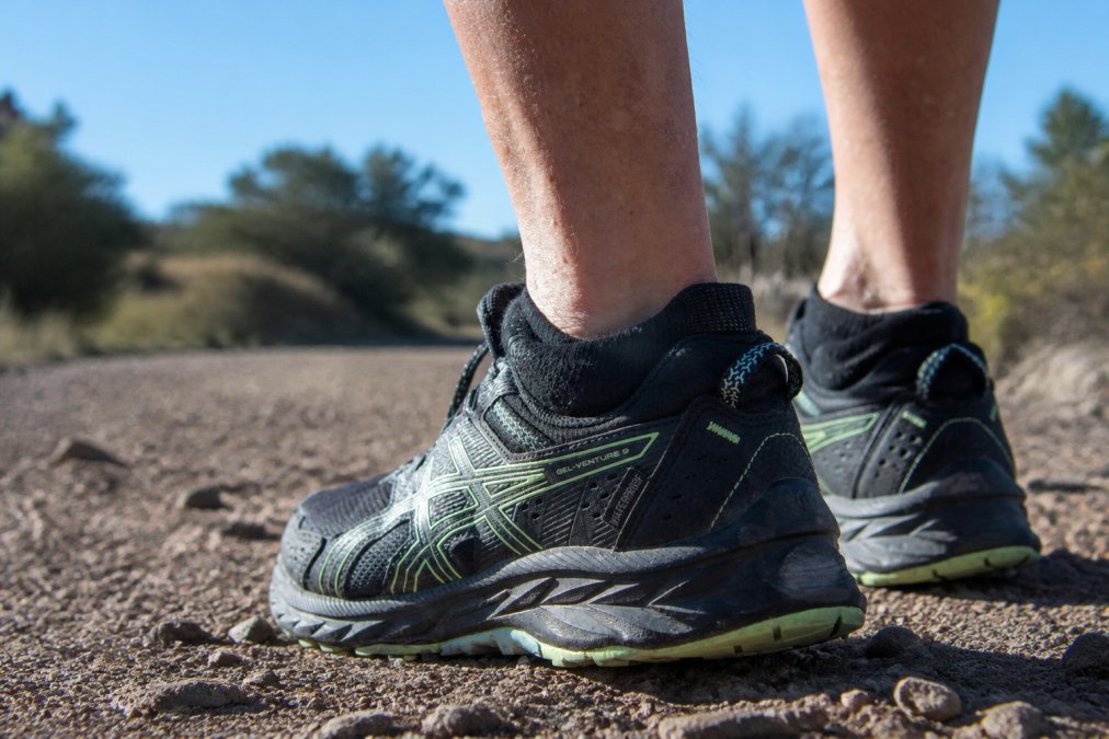 Close-up of runner's feet during a long-distance trail run on rocky terrain, showing proper sock fit with moisture-wicking fabric visible and debris barrier preventing dirt entry