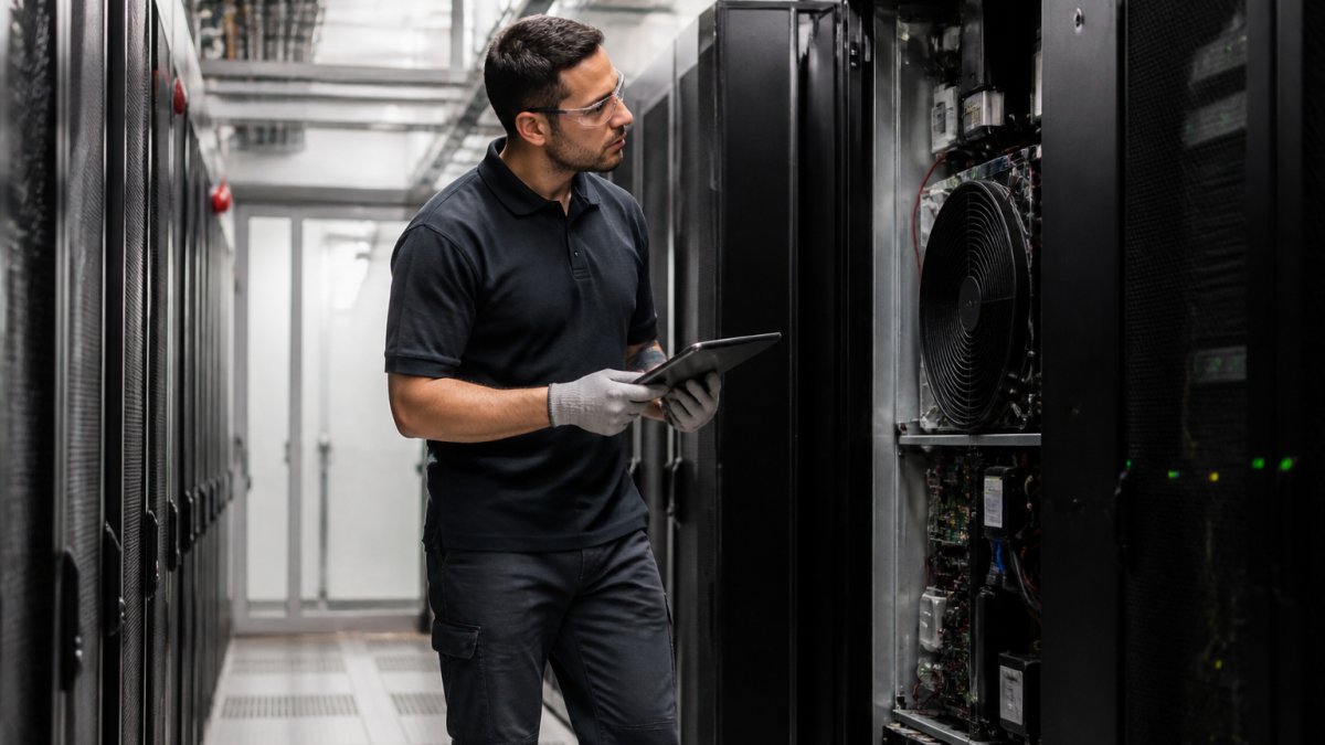 A technician in safety glasses and gloves inspecting the open front panel of a precision cooling unit in a data centre aisle, tablet in hand, with server racks visible behind