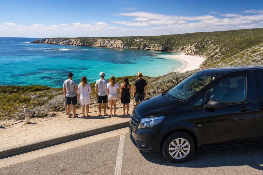 Scenic coastal landscape with turquoise waters, white sand beach, and small group of tourists enjoying the view with professional transport vehicle parked nearby, natural daylight