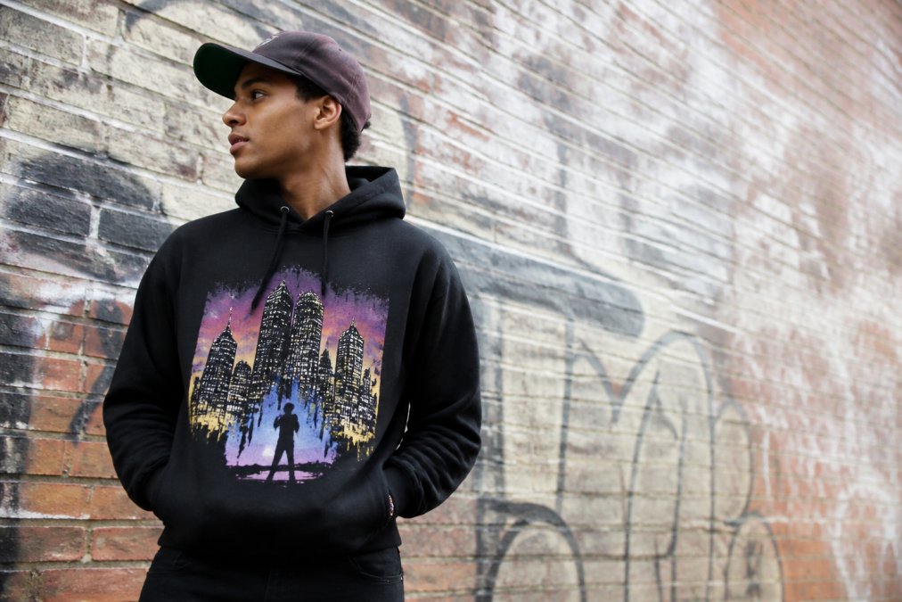 Eine junge Person in einer modernen Stadt trägt einen grafischen Hoodie und eine auffällige Kappe und wird vor einer Backsteinmauer mit leichter Graffiti, im Streetwear-Stil und bei natürlichem Tageslicht fotografiert.