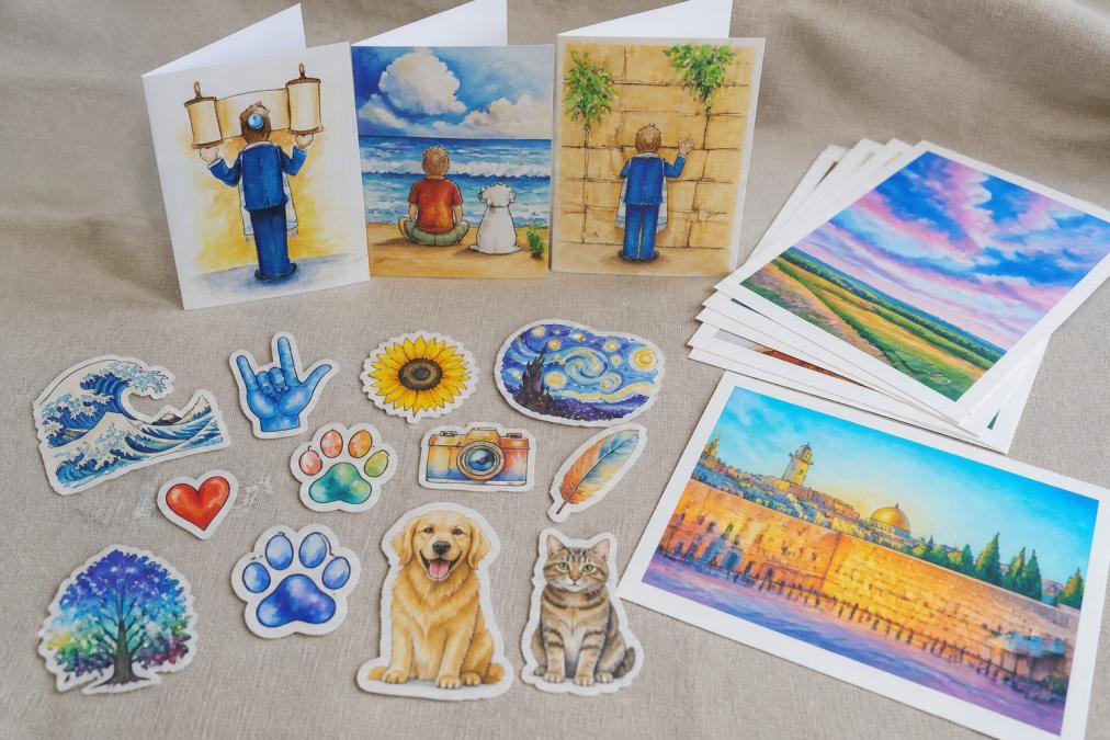 Arranged display of handmade greeting cards with vibrant watercolor designs, vinyl stickers in various colors and themes, and original art prints laid out on a neutral linen surface, showing the range of artistic styles and product types