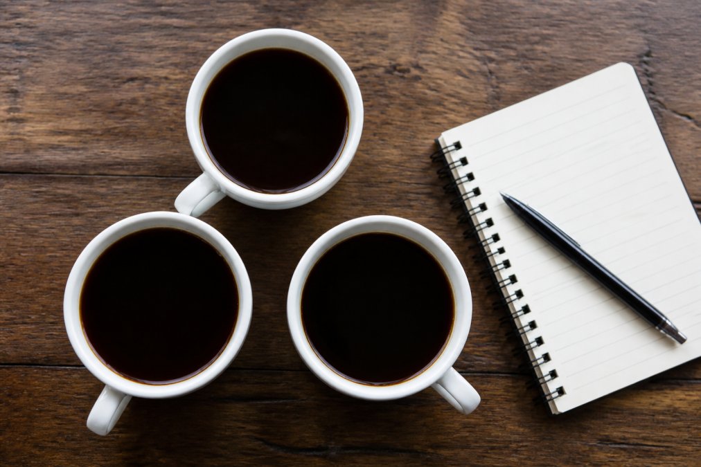 Overhead shot of three white coffee cups on a wooden table, each filled with dark coffee, with a notebook and pen beside them for tasting notes