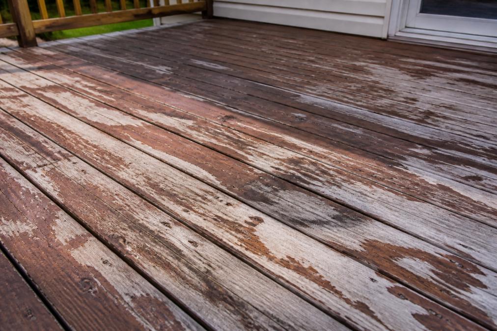 Weathered wooden residential deck showing peeling and fading oil-based stain with visible deterioration, bare patches exposing raw wood, and water damage marks under natural afternoon sunlight