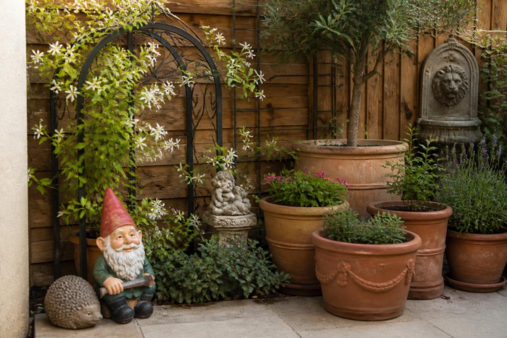 A small patio corner featuring a ceramic garden gnome positioned near a rustic wrought iron arch with climbing jasmine, terracotta pots in graduating sizes creating layers, and ornamental statues arranged to establish a focal point in a confined outdoor space