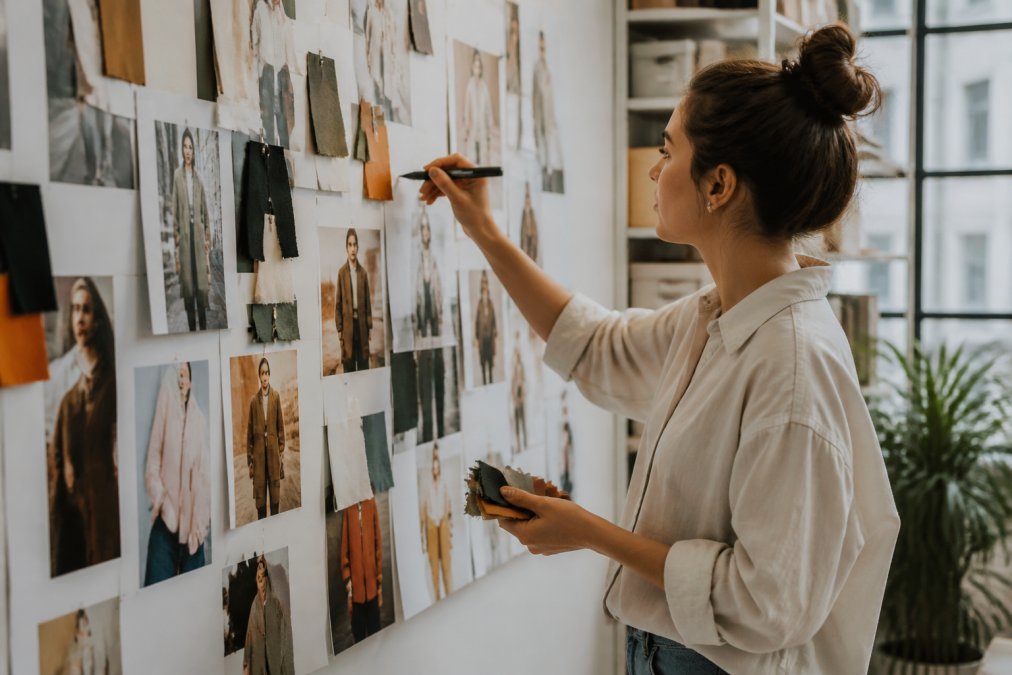 Person arranging printed fashion inspiration images, fabric color swatches, and style reference photos on a white wall or cork board while holding a marker to annotate style notes