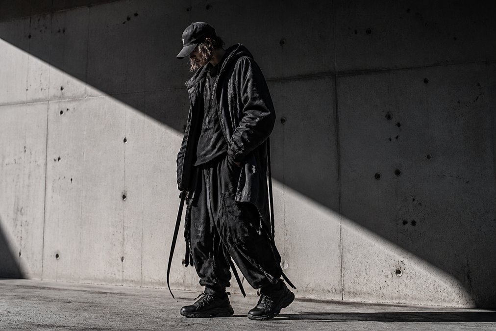 Person wearing layered dystopian streetwear with deconstructed silhouettes, oversized straps, and industrial hardware details against an urban concrete wall with dramatic shadow lighting