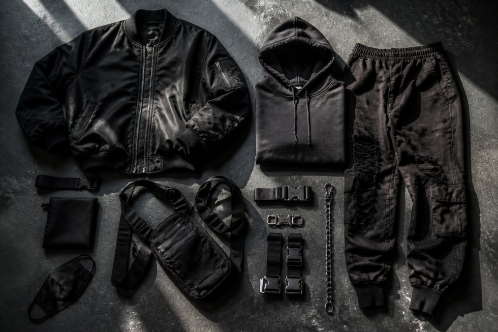 Flat lay arrangement of futuristic fashion essentials including oversized bomber jacket, monochromatic hoodie, distressed joggers, industrial straps, and hardware accessories arranged on dark textured surface with dramatic side lighting