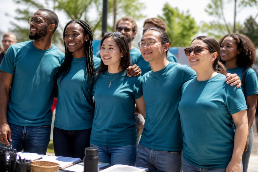 Diverse group of activists at a grassroots organizing event wearing matching democracy t-shirts, standing together in sunlight, showing solidarity and collective strength