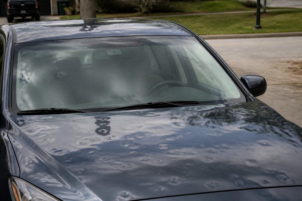Vehicle with visible dent damage across the hood and roof from hail impact, parked outdoors in natural daylight, showing the scattered pattern of dent damage typical of hail storms