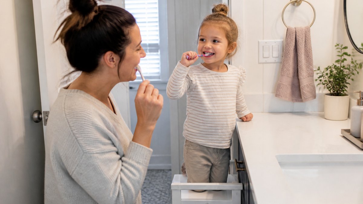 A parent and young child brushing their teeth together in a bright bathroom, both smiling, with a step stool and two toothbrushes visible