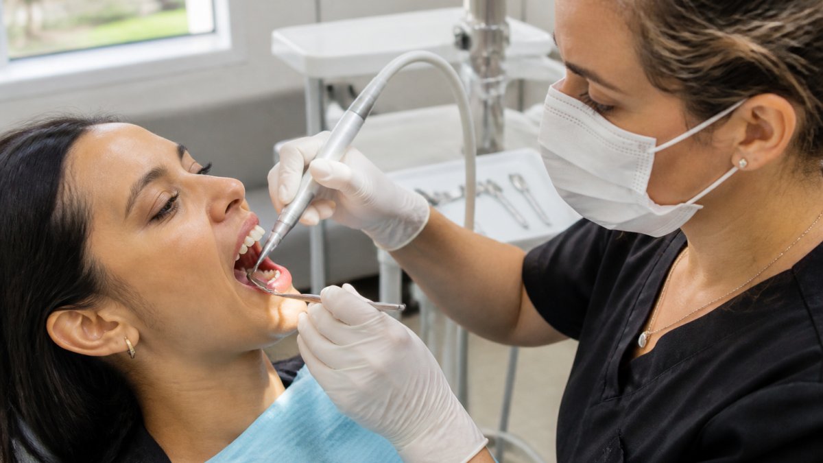 Professional dental hygienist performing scaling and root planing procedure on patient's lower teeth using ultrasonic scaler in modern dental office with natural lighting