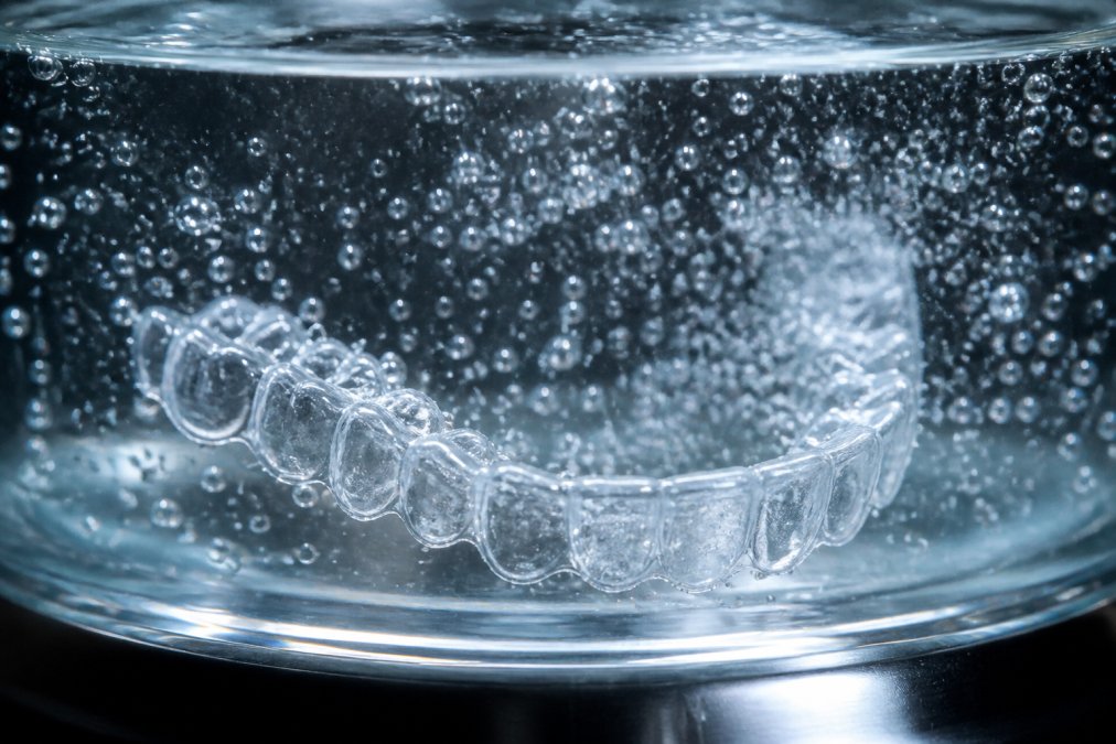 Nahaufnahme einer Zahnschiene in klarem Wasser mit sichtbaren Mikrobläschen und Schallwellen-Effekten, zeigt den Reinigungsprozess in Aktion