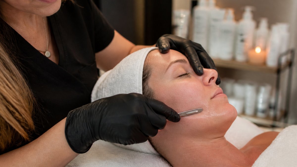Close-up of an esthetician in gloves using a sterile scalpel blade along a client's jawline in a calm, softly lit treatment room, client relaxed with hair wrapped in a towel