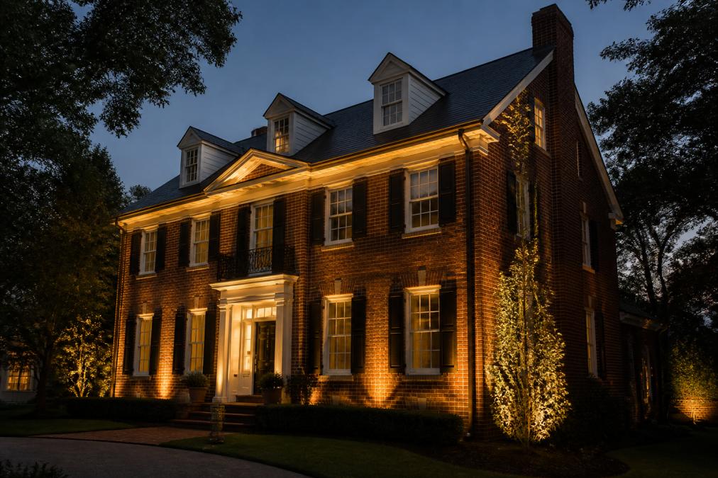 Brick colonial home photographed at dusk with warm uplighting illuminating the facade and foundation, showing the contrast between lit and unlit architectural details