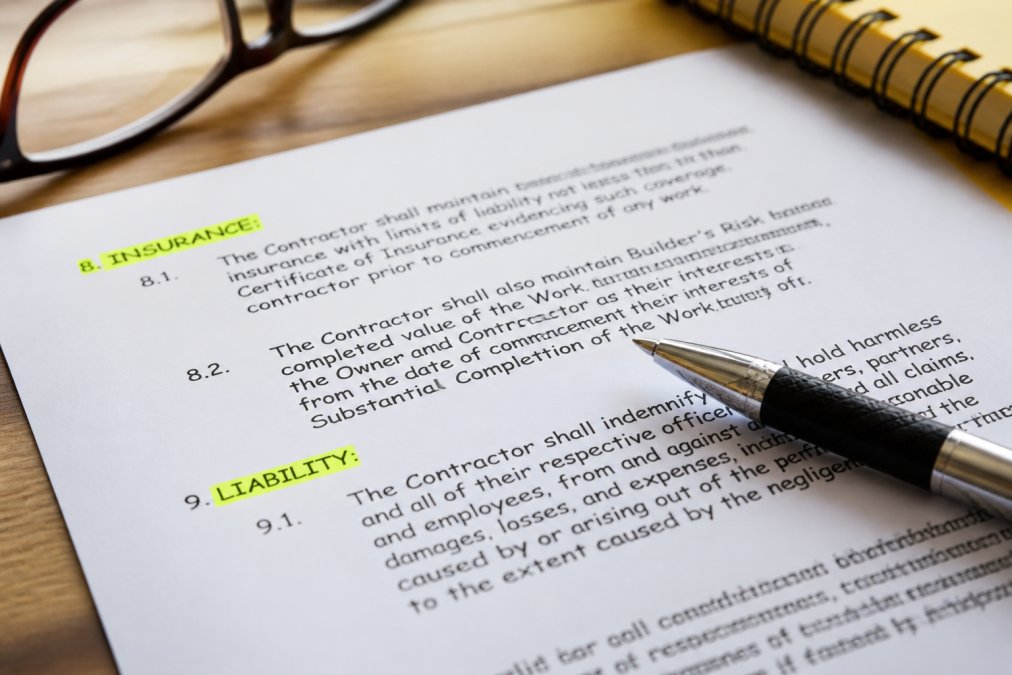 Close-up of a construction contract document with sections highlighted, showing insurance requirements and liability clauses, pen resting on the page, professional office setting
