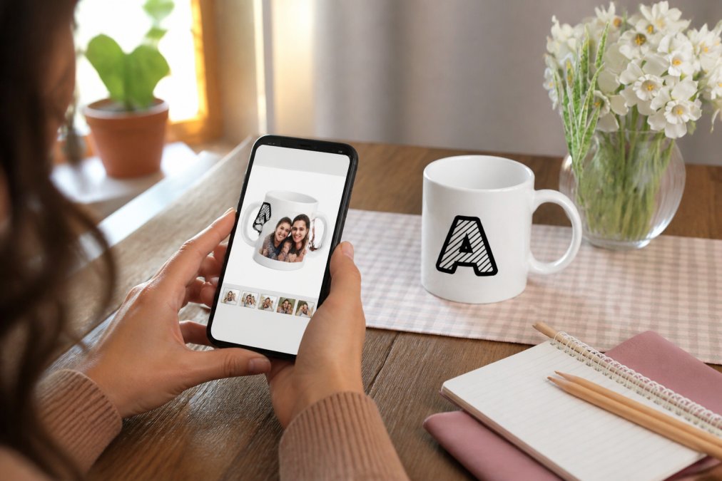 Person holding a smartphone displaying a custom mug design tool interface with a family photo positioned on a white ceramic mug preview, sitting at a wooden desk with natural sunlight streaming through