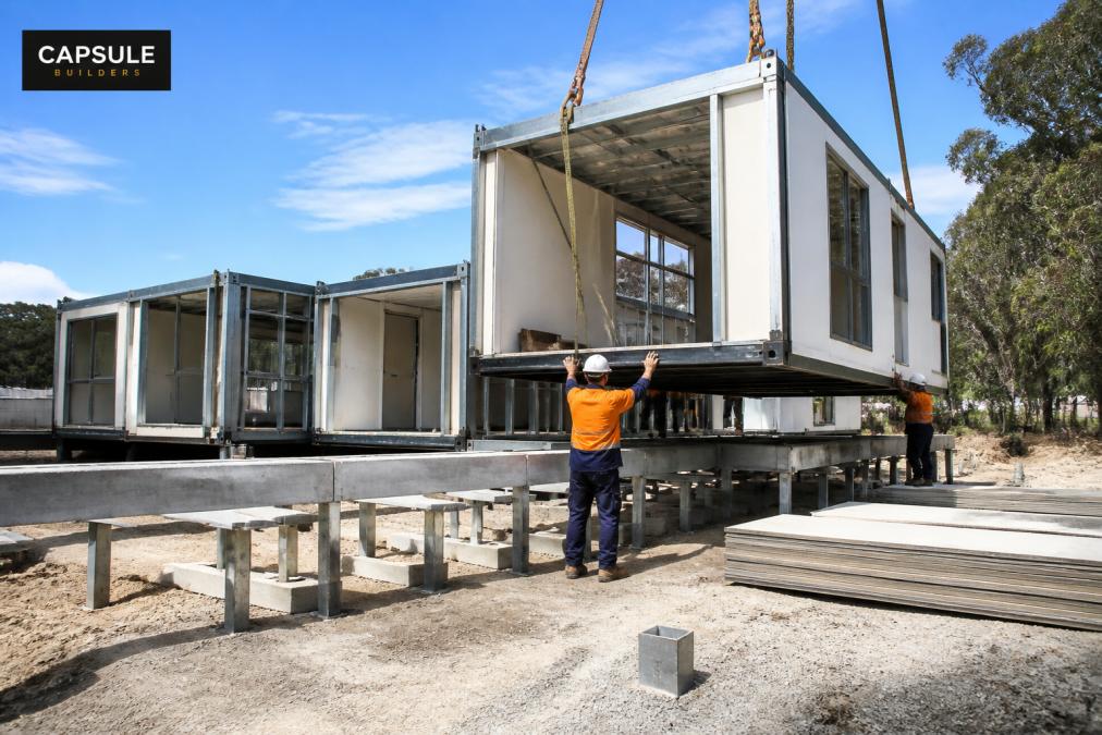 Steel-framed modular sections being assembled on residential site, showing precision construction with workers positioning modules, multiple fabricated sections visible, daytime site lighting