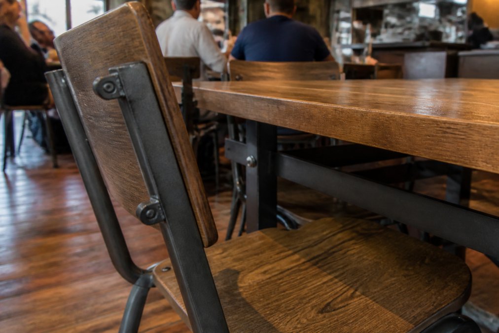 Close-up of a commercial-grade restaurant chair and table in a busy dining room, showing reinforced metal frame joints and a solid wood tabletop with visible wear-resistant finish, diners seated in the background