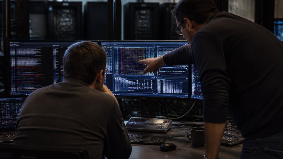 A security analyst in a dimly lit operations center reviewing multiple monitors displaying AI agent execution logs and anomaly detection alerts, with a colleague pointing at a specific data pattern on screen