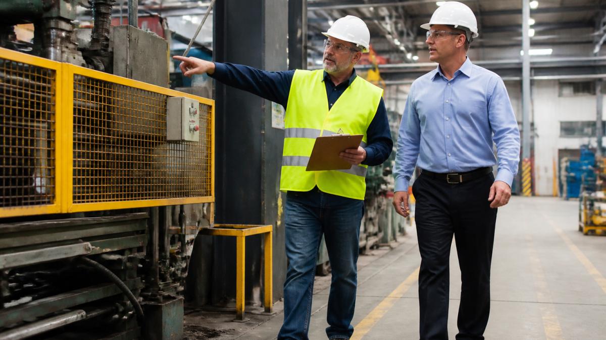 A safety manager in a hard hat and safety vest walking a manufacturing floor with a clipboard, pointing out a machine guard to a plant supervisor