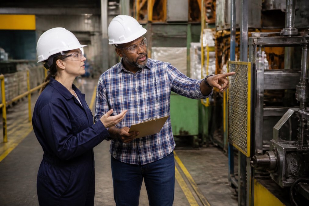 Safety manager and worker conducting a walk-through inspection on manufacturing floor, using clipboard to document equipment hazards and pointing out potential danger zones