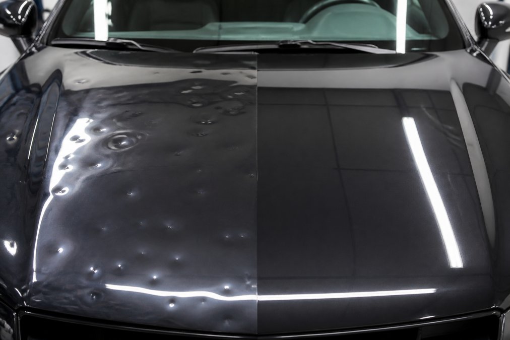 Close-up view of a car hood showing a hail-dented surface on the left half and a smooth, flawless factory paint finish on the right half after PDR repair, photographed under bright studio lighting to highlight the contrast