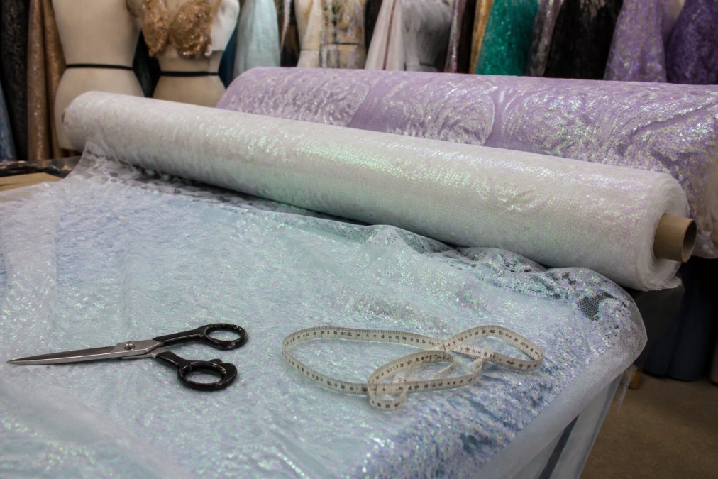 A professional designer's workspace with rolls of iridescent sequin stretch mesh fabric in damask patterns, scissors and a measuring tape on a cutting table