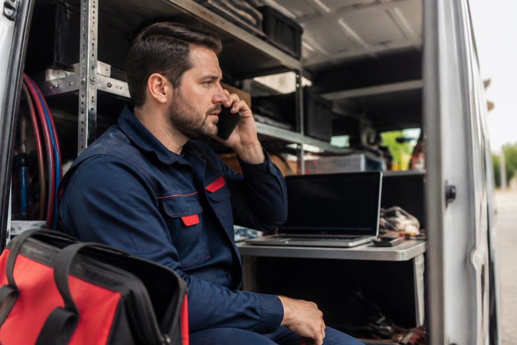 An HVAC technician in a branded uniform answering a phone call inside a service van, with a laptop and dispatch software visible in the background