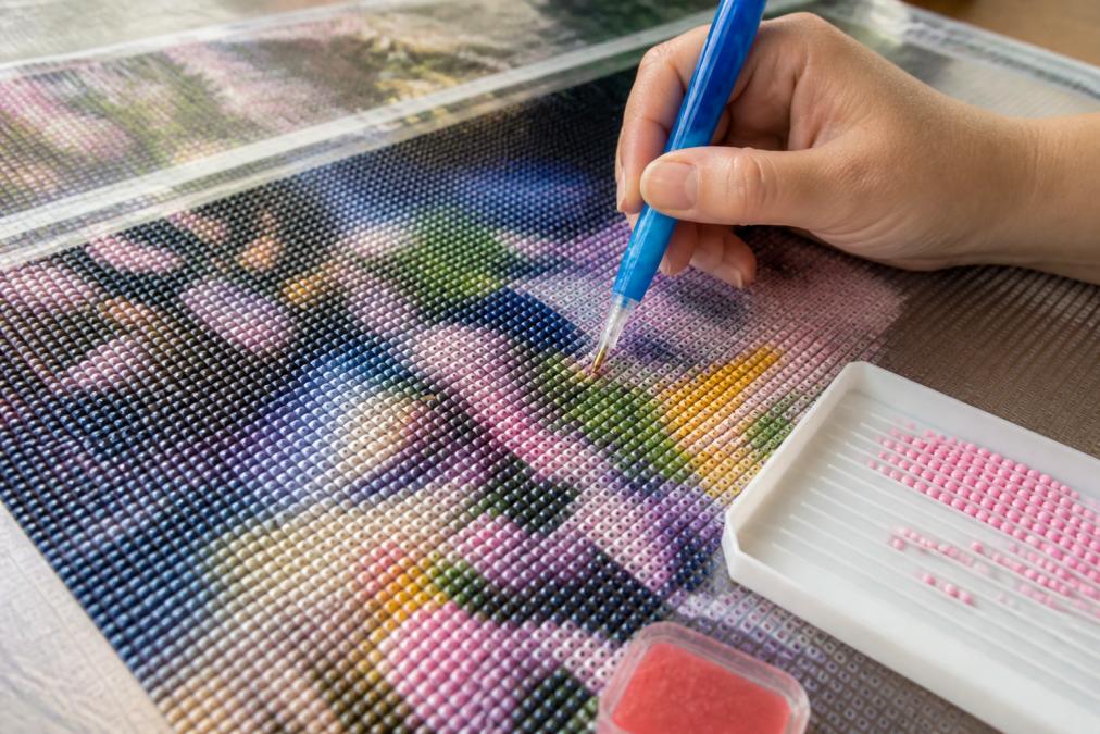 Close-up of hands placing colorful resin drills on a diamond painting canvas with an applicator tool and wax caddy visible on a work surface