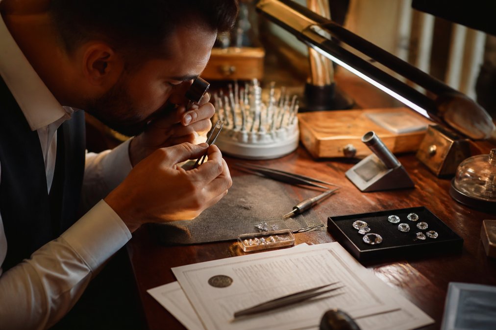 A professional jeweler with magnifying loupe examining a loose diamond under bright, focused lighting at a jeweler's workbench, with gemological tools and certification documents visible